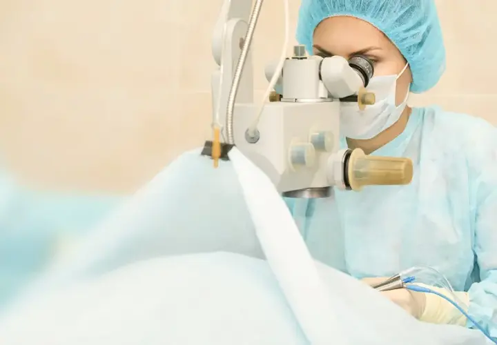 Chirurg w masce i czepku wykonuje operację zaćmy, używając mikroskopu i precyzyjnych narzędzi.