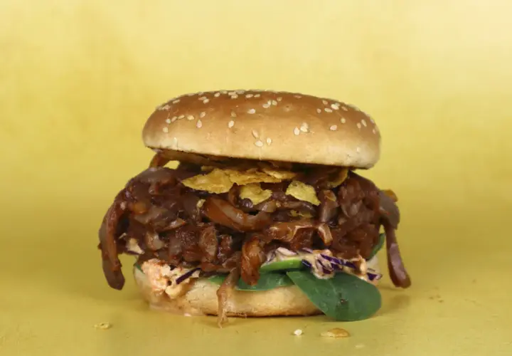Hamburguesa con pulled pork vegano, cebolla caramelizada y crujientes patatas fritas sobre fondo amarillo.