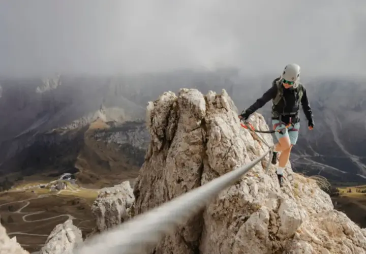 Kobieta na via ferrata polska pokonuje skalną grań, w tle górzysty krajobraz i kręta droga.