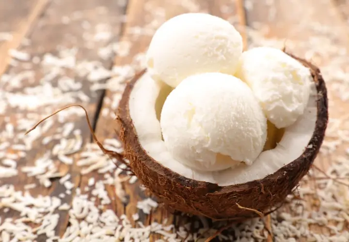 Tre palline di cremoso gelato al cocco servite in una noce di cocco su un tavolo di legno con scaglie di cocco sparse.