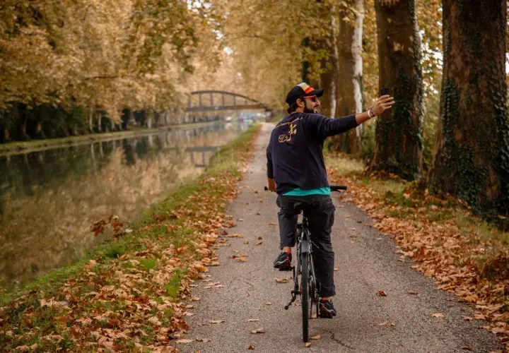 Hombre en bicicleta toma selfie en el Canal du Midi, rodeado de árboles otoñales y hojas caídas.