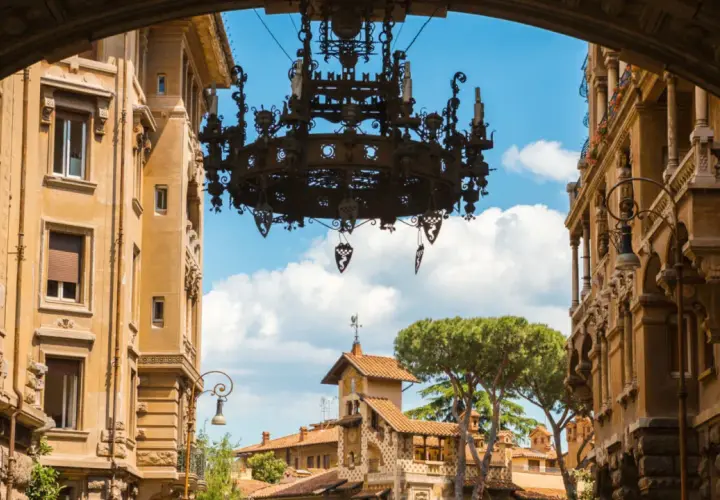Un imponente lampadario domina la vista del quartiere Coppedè, incorniciato da palazzi storici e un cielo azzurro con nuvole bianche.