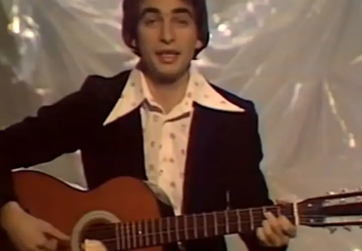 Mężczyzna w garniturze i wzorzystej koszuli gra na gitarze. W tle widać materiał, na którym **ciągle pada tekst**.