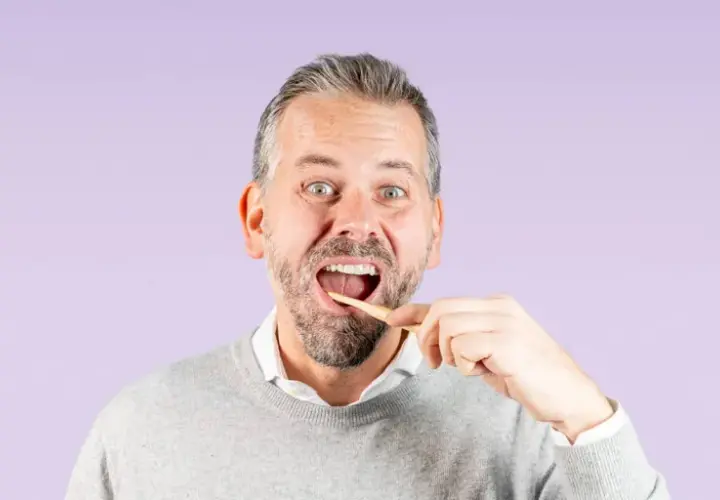 Uomo con barba che si lava i denti con uno spazzolino di legno, un gesto quotidiano per una corretta igiene dentale.