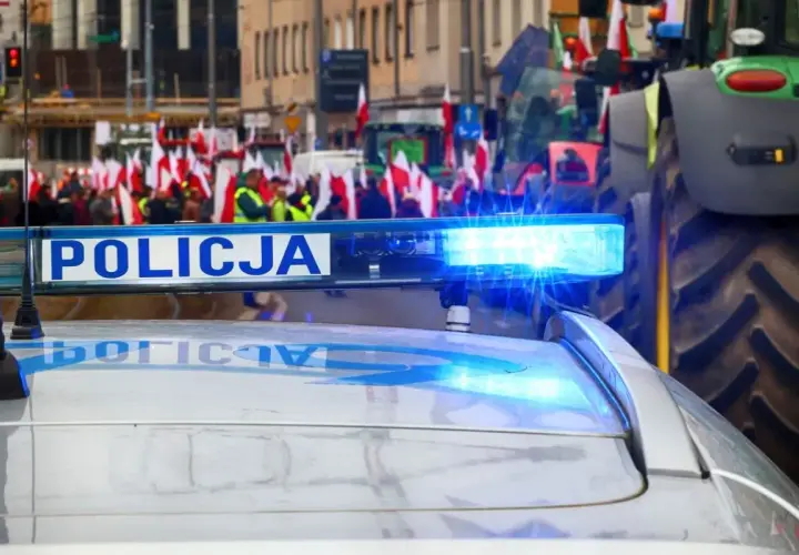 Policyjny radiowóz z włączonym sygnałem świetlnym na tle protestu rolników z flagami. Czy to jego jaka odprawa po 20 latach w policji?