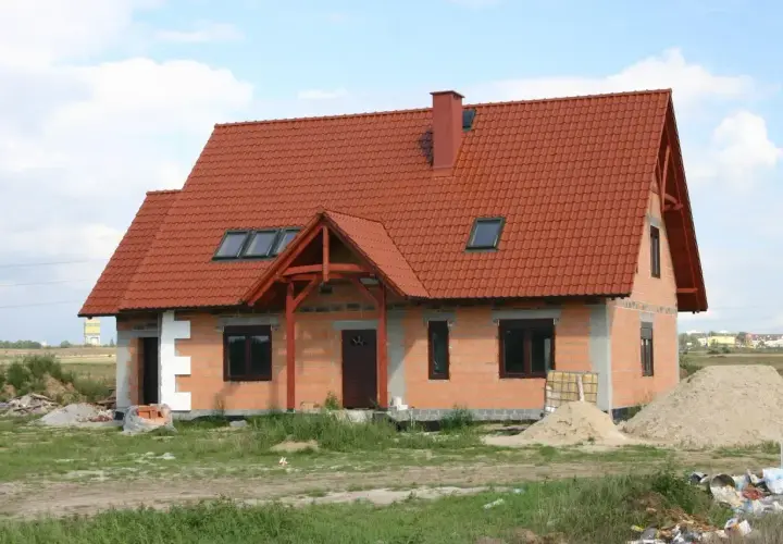 Obecny sposób zagospodarowania terenu inwestycji to budowa domu z czerwonym dachem, otoczonego materiałami budowlanymi i zielenią.