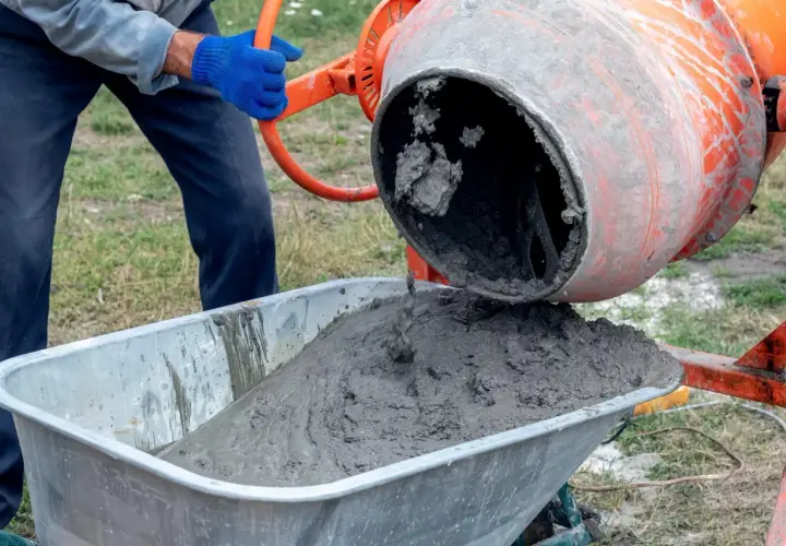 Béton frais coulant d'une bétonnière orange dans une brouette. Un ouvrier en gants bleus manipule la manivelle pour déverser le mortier béton.