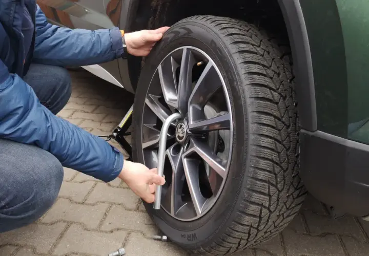 Osoba w niebieskiej kurtce i jeansach dokonuje wymiany koła w samochodzie, używając podnośnika i klucza.