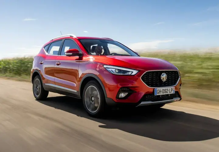 MG ZS en prueba, rojo brillante, circulando por un camino rural con campos verdes y cielo azul.