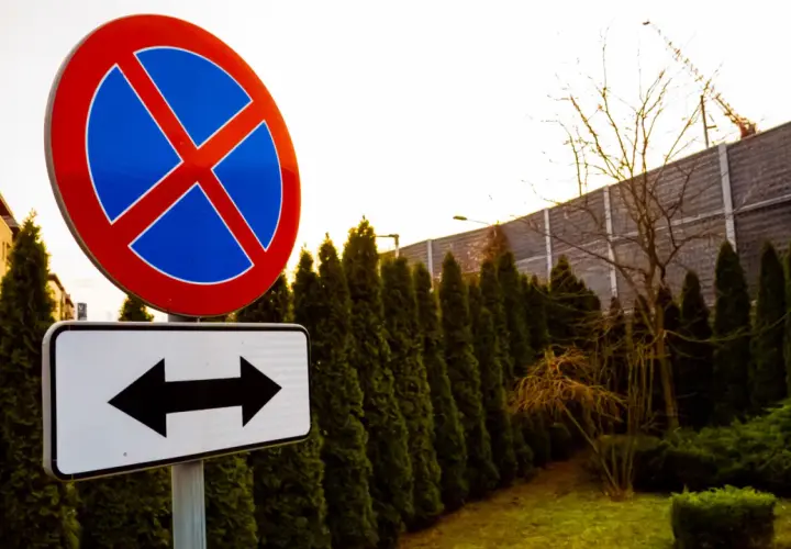 Znak zakaz zatrzymywania z tabliczką wskazującą kierunki. W tle budynek i żywopłot.