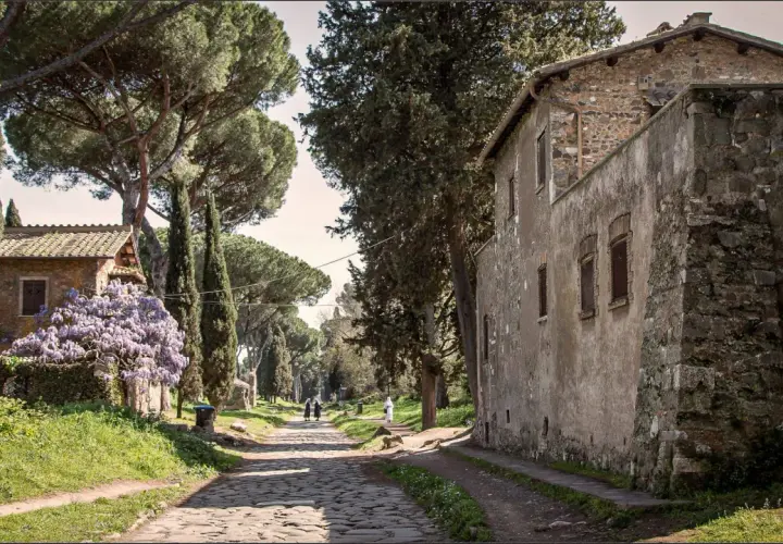 Une ancienne voie romaine à Rome, bordée de cyprès et de pins parasols, avec des maisons en pierre et des glycines en fleurs.