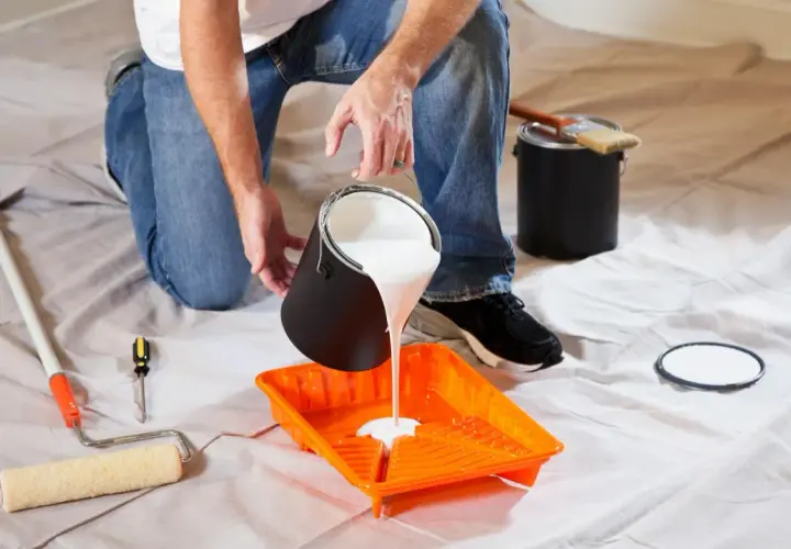 Uomo versa vernice bianca in una vaschetta arancione, pronto per pitturare soffitto con pennello. Attrezzi da pittura a portata di mano.