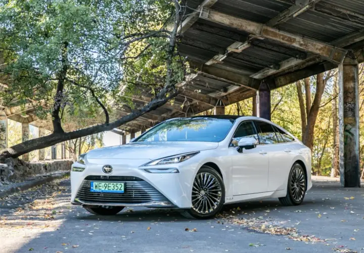 Biała Toyota Mirai, samochód przyszłości, zaparkowana pod zadaszeniem z opadłymi liśćmi wokół.