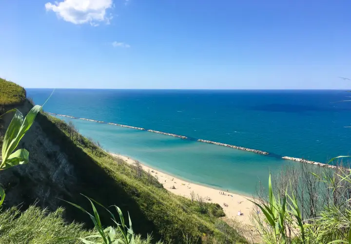 Vista panoramica della spiaggia di Pesaro con mare azzurro e scogliera verde. La focara è un ricordo lontano, ma la bellezza del paesaggio resta.