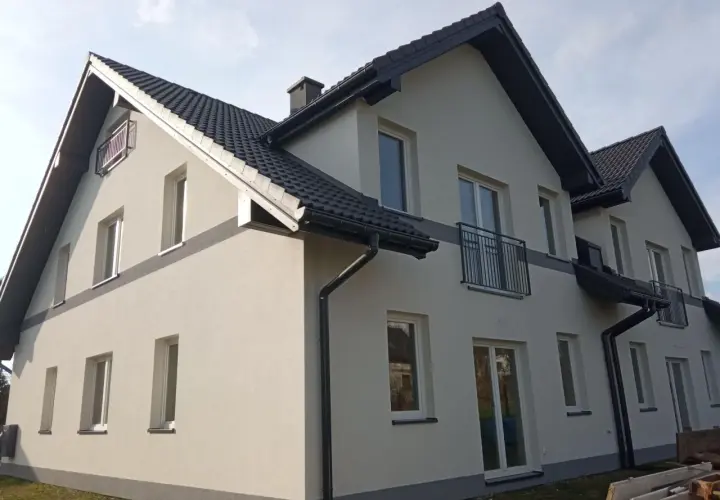 Nowy dom z jasną elewacją i ciemnym dachem. Wokół zielona rząska trawy, a na ścianach liczne okna i balkony.