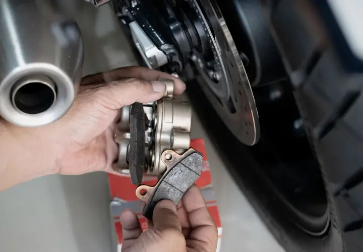 Mani che effettuano il cambio pastiglie freni di una moto, mostrando le vecchie pastiglie usurate e il nuovo set pronto per l'installazione.