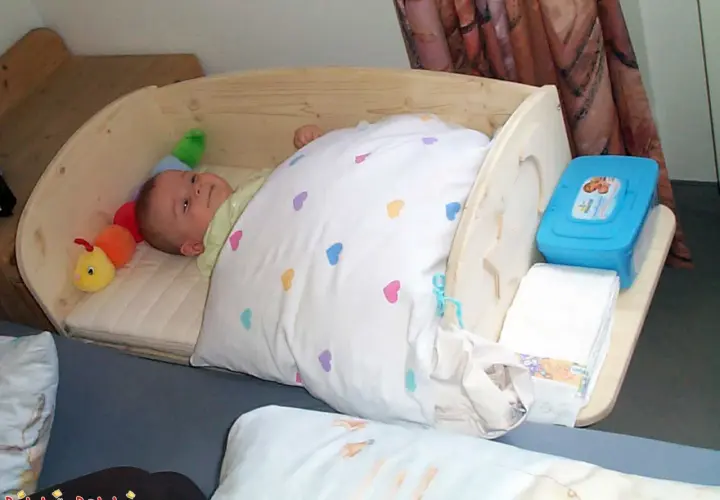 Ein süßes Baby schläft in einem selbstgebauten Babybett aus Holz. Neben ihm liegt ein bunter Raupen-Greifling. Windeln und Feuchttücher sind griffbereit.