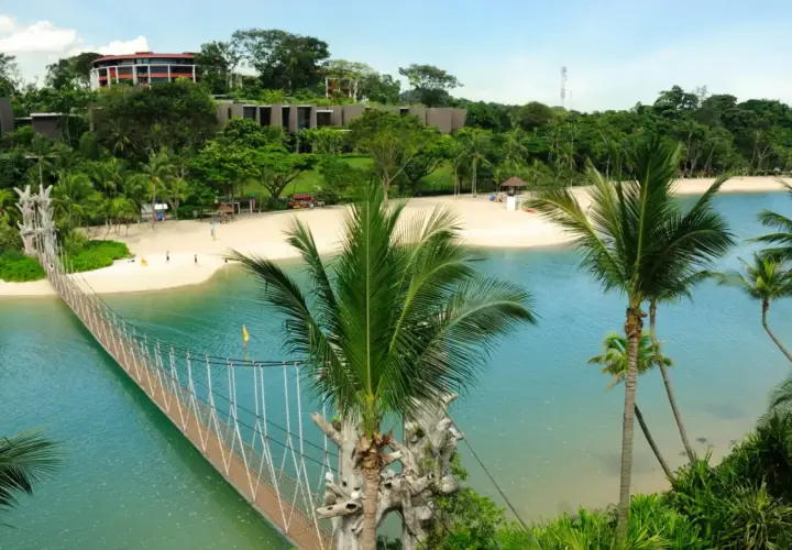 Hängende Brücke über türkisfarbenem Wasser führt zu einem sonnigen singapur strand mit Palmen und Resorts im Hintergrund.