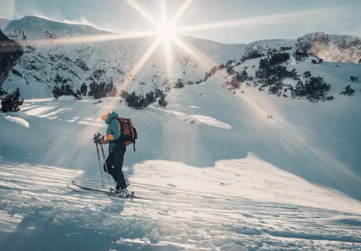 Skitouring co to? Narciarz z plecakiem wspina się po ośnieżonym stoku, słońce przebija się przez chmury, tworząc magiczną atmosferę.
