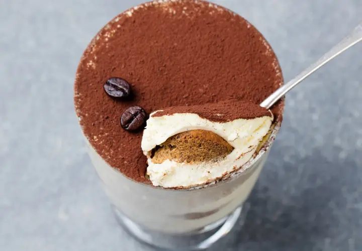 Un goloso tiramisù giallo zafferano, cosparso di cacao e chicchi di caffè, pronto per essere gustato con un cucchiaino.