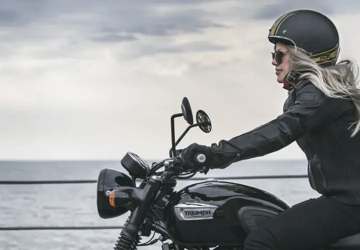 Mujer rubia en moto Triumph negra, con casco y chaqueta de cuero, disfrutando del paisaje. Las motos Triumph se fabrican con pasión.