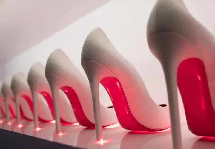 Rząd eleganckich, jasnych szpilek z czerwonymi podeszwami, przypominających kultowe Louboutiny, lśni w blasku neonowego światła.