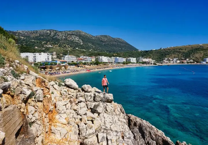 Mann steht auf Felsen mit Blick auf eine Bucht mit Strand und Hotels. Perfekt, um zu wissen, wo in Albanien Urlaub machen!