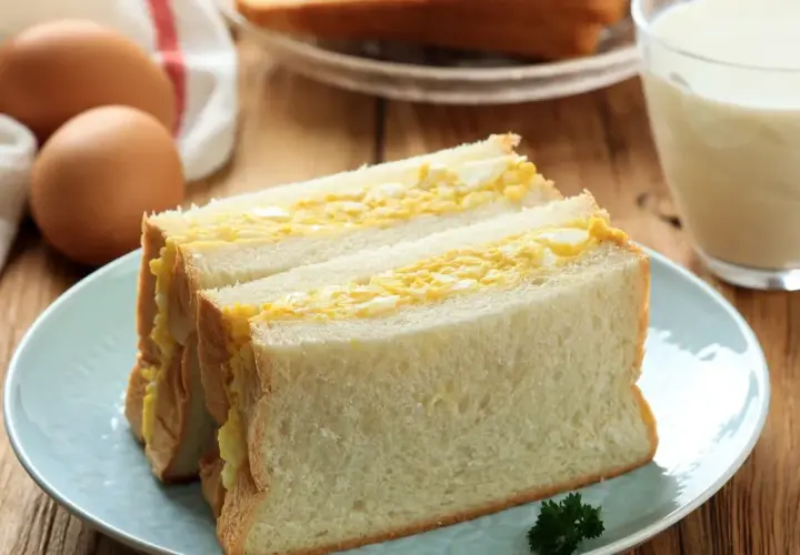Un délicieux sandwich aux œufs, garni de pain blanc moelleux, servi sur une assiette bleue avec un verre de lait.