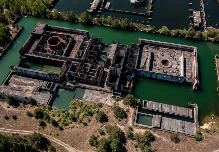 Niedokończona elektrownia atomowa Żarnowiec, z betonowymi konstrukcjami zanurzonymi w zielonej wodzie, otoczona drzewami i dziką roślinnością.