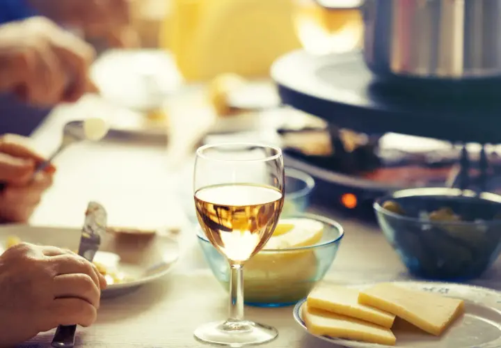 Un verre de vin blanc sec ou moelleux accompagne une raclette conviviale. Des morceaux de fromage et des cornichons sont prêts à être dégustés.