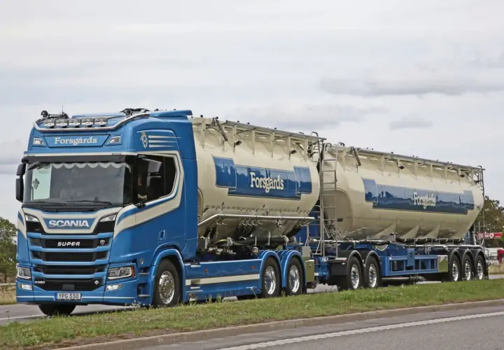 Ein blauer Scania-Sattelzug mit zwei Siloanhängern, beladen für Forsgårds, fährt auf einer schwedischen Straße. Das Tempolimit in Schweden ist zu beachten.