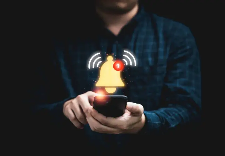 Dłoń trzymająca smartfon, nad którym unosi się złoty dzwonek z powiadomieniem.