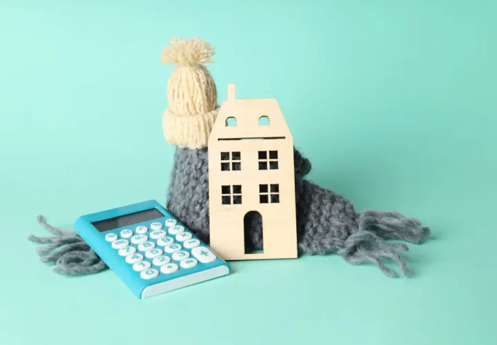 Casa de madera abrigada con bufanda y gorro, junto a calculadora. Simboliza el cálculo del coeficiente de rendimiento energético para el hogar.