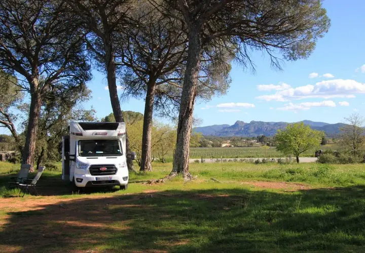 Un camping-car blanc est garé sous des pins, avec des chaises pliantes à côté. Un paysage de vignobles et de montagnes s'étend au loin. Un guide aire camping car idéal pour l'aventure.