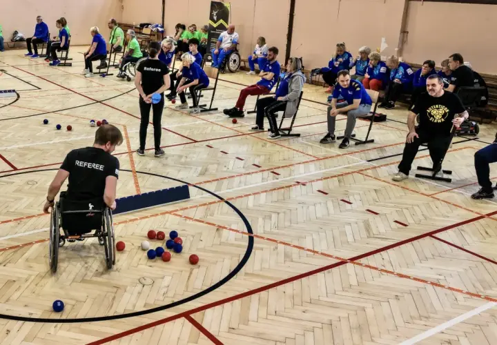 Zawodnicy grają w boccię, sportową grę precyzyjną. Na boisku widać kolorowe kule i zawodnika na wózku inwalidzkim.