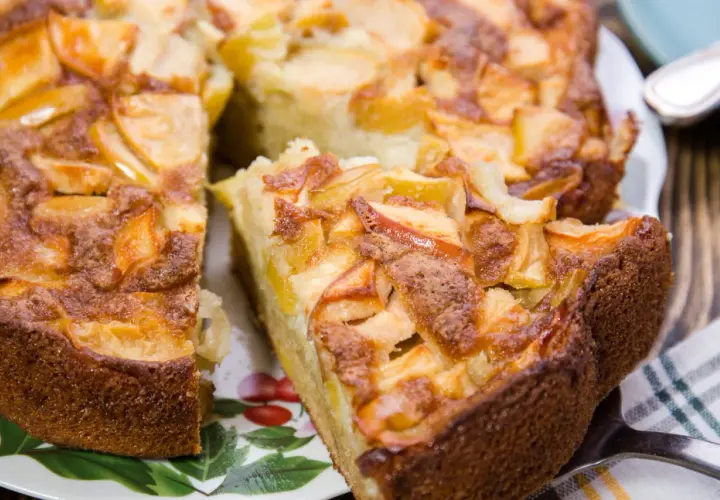 Un délicieux gâteau aux pommes, doré et moelleux, avec des morceaux de fruits caramélisés sur le dessus. Une part est déjà coupée, prête à être dégustée.