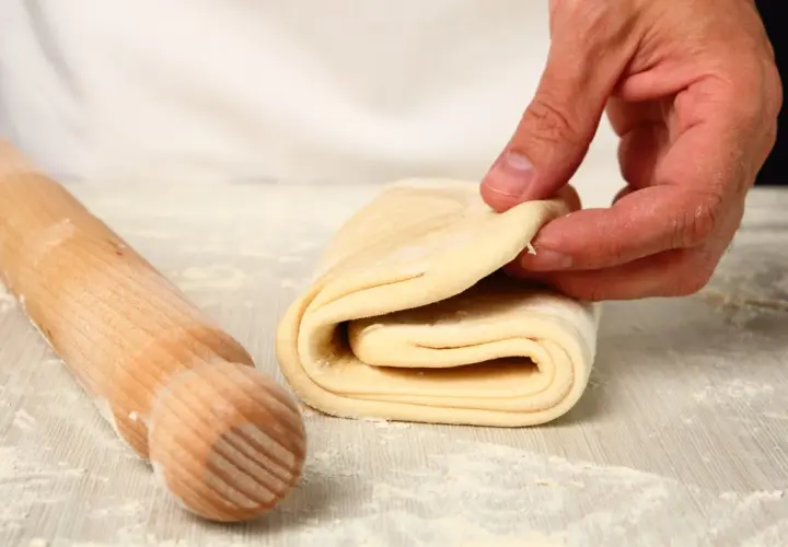 Mani che piegano pasta sfoglia, pronta per diventare pasta fatta in casa. Un mattarello è accanto, pronto per il prossimo passaggio.