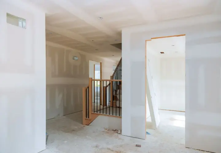 Interno di una casa in costruzione con pareti in cartongesso grezzo, una scala in legno e metallo e un'apertura verso un'altra stanza.