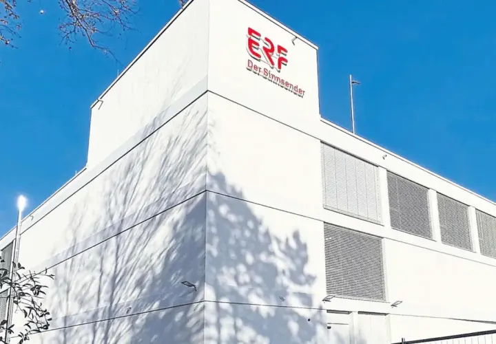 Das Gebäude von ERF, dem Sinn-Sender, unter blauem Himmel. Schatten von Bäumen tanzen auf der weißen Fassade.