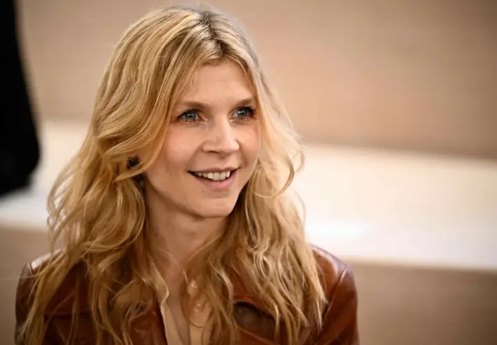Uśmiechnięta Clémence Poésy z falującymi blond włosami i niebieskimi oczami, ubrana w brązową skórzaną kurtkę.