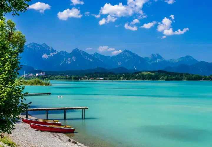 Türkisblaues Wasser am Badesee Bayern, mit roten Ruderbooten am Kiesstrand und majestätischen Alpen im Hintergrund.