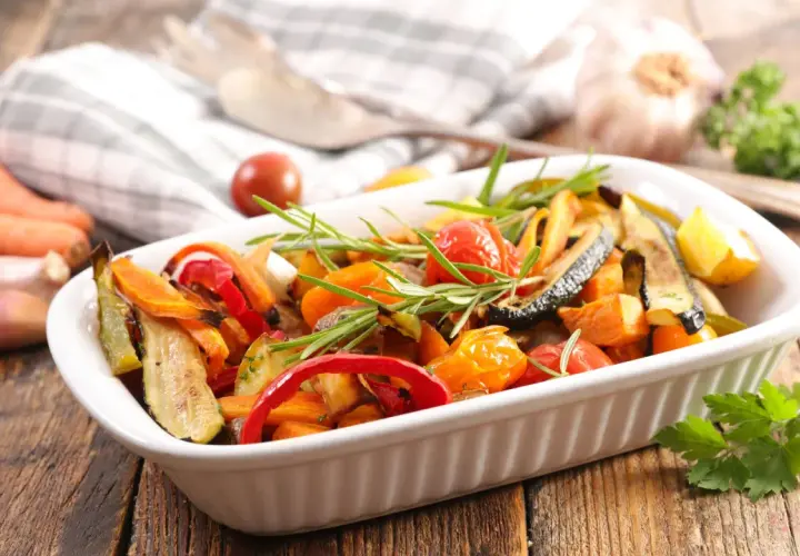 Un plato blanco lleno de coloridas verduras al horno, con romero fresco y tomates cherry.