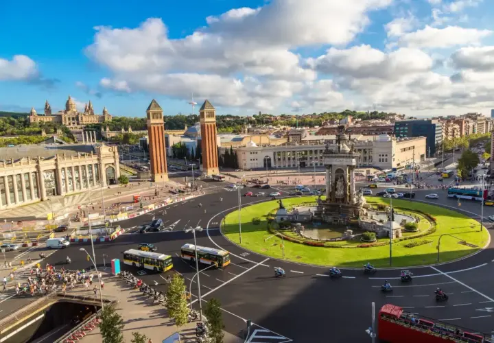 Plac Hiszpański w Barcelonie, z dwoma wieżami w tle i fontanną pośrodku. Ruch uliczny, autobusy i motocykle tworzą tętniącą życiem scenę w tym pięknym, **starym mieście Barcelona**.