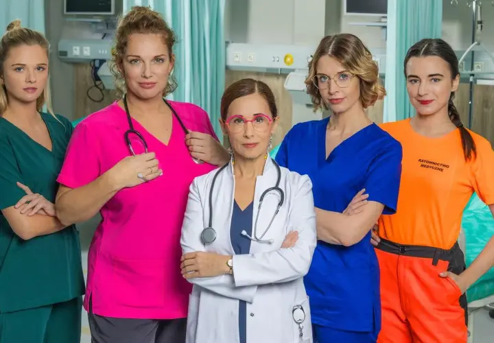Pięć kobiet w strojach medycznych, obsada serialu "Szpital św. Anny", pozuje do zdjęcia w szpitalnej sali.