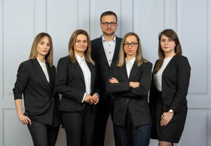 Pięć osób w garniturach, zespół prawników, stoi przed jasną ścianą.