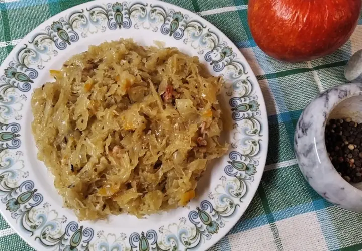 Na talerzu z ozdobnym wzorem znajduje się porcja pysznej, **kapusty kiszonej gotowanej**. Obok jabłko i moździerz z pieprzem.