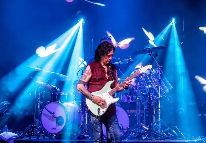 Mężczyzna gra na gitarze elektrycznej na scenie, w tle perkusja i niebieskie światła z motylami.