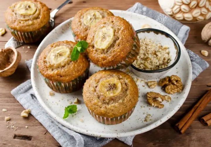 Pięć pysznych muffinek bananowych z plasterkiem banana na wierzchu, udekorowane miętą. Obok miseczka z orzechami i laska cynamonu.