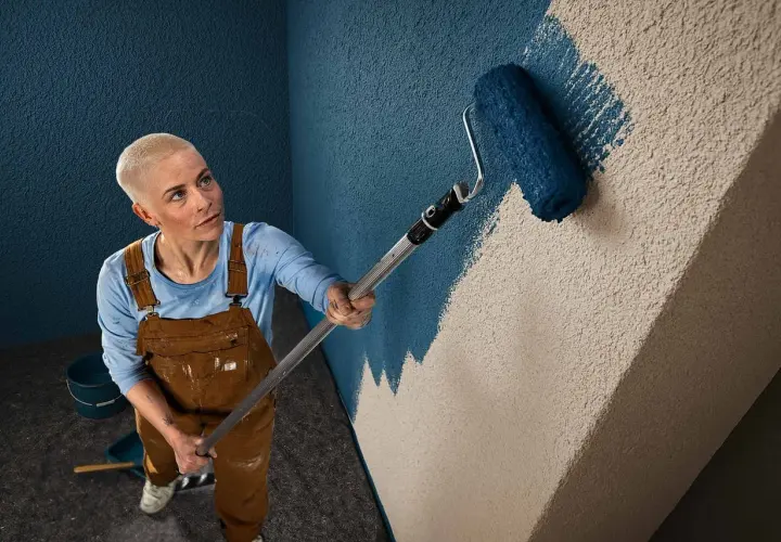 Frau mit Kurzhaarschnitt streicht mit blauem Roller eine Wand mit Rauputz.