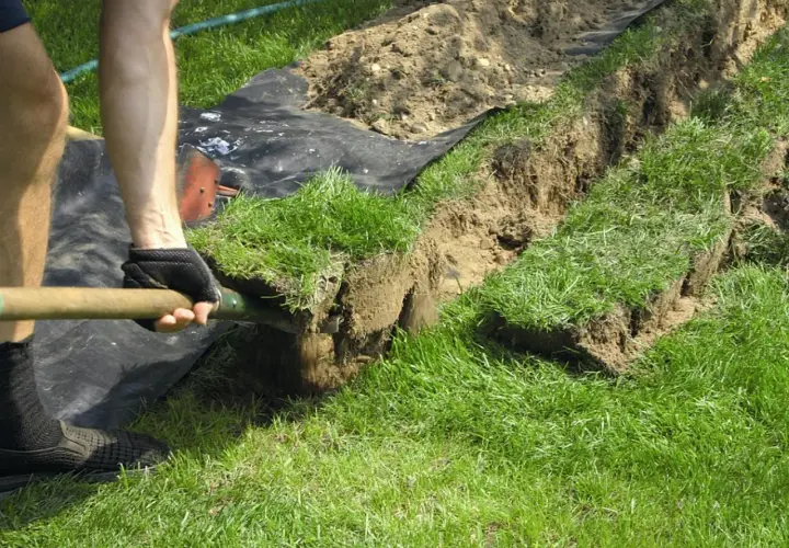 Osoba w rękawiczkach kopie łopatą rów, wycinając darnie. Prace ogrodowe w pełnym słońcu.
